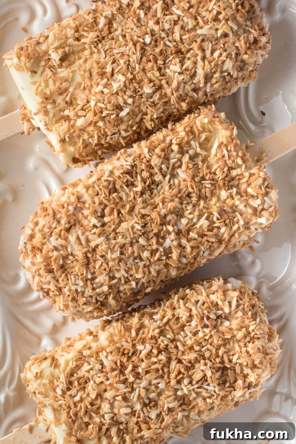 homemade coconut ice cream bars coated in toasted coconut resting on a white textured plate. 