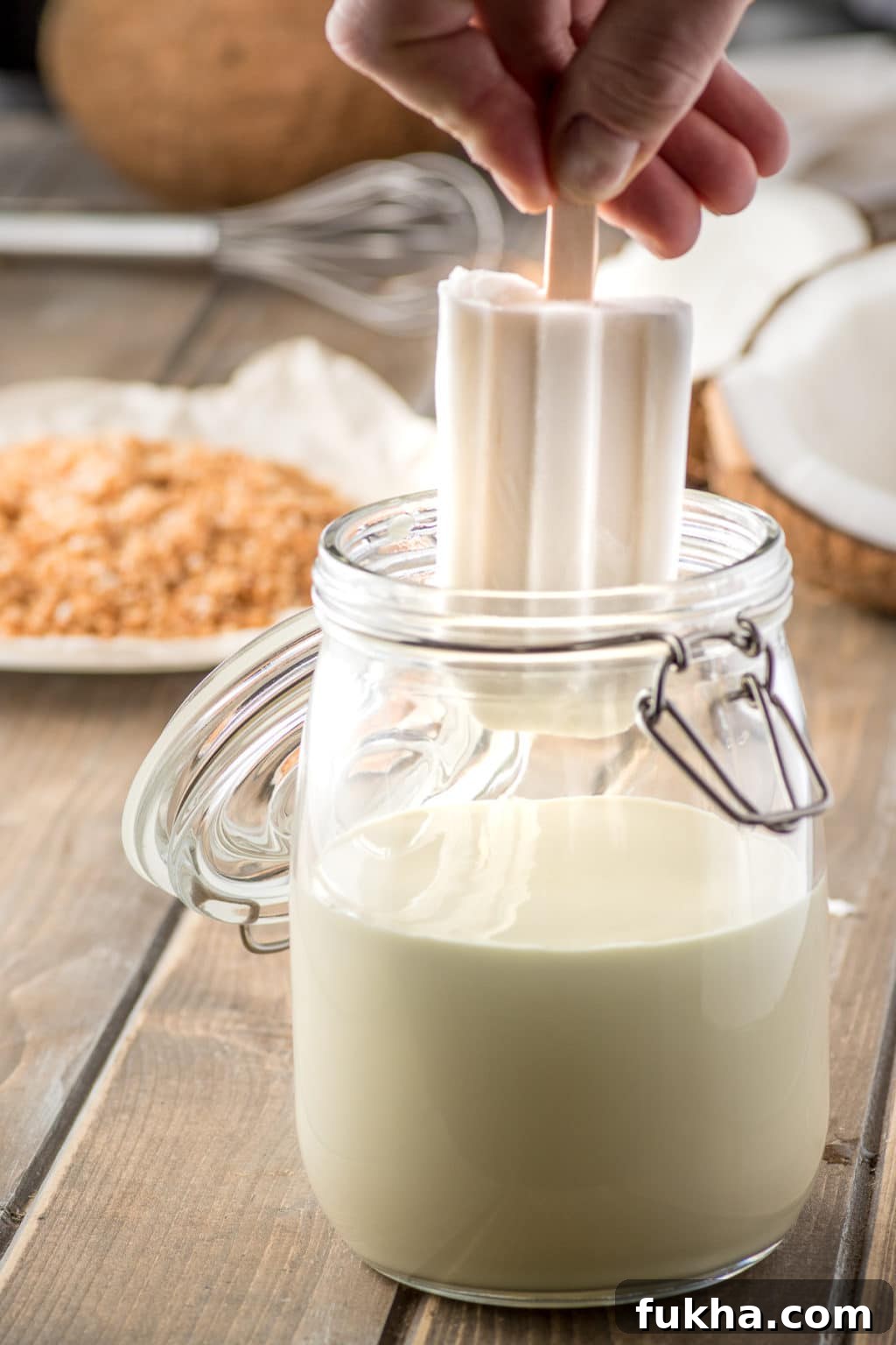 a glass jar filled with white chocolate coating with a white coconut popsicle being dipped 