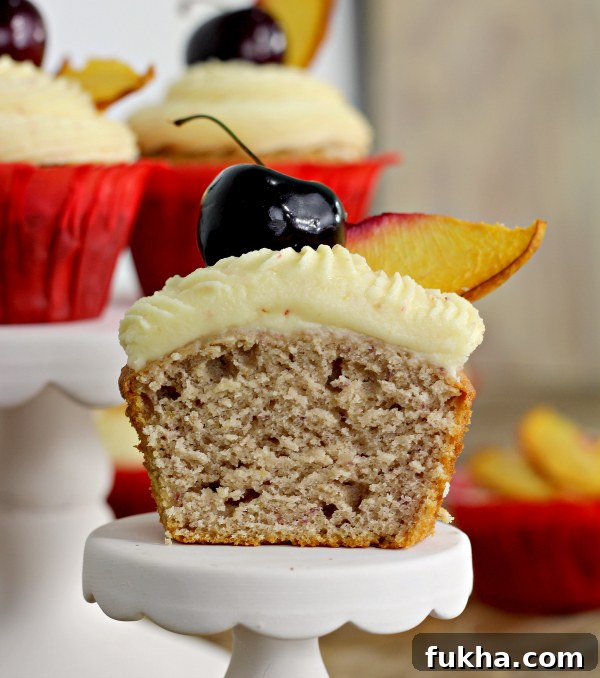 a peach cupcake cut in half with cherry and peach slice on top of frosting. The cupcake is sitting on a cupcake stand 
