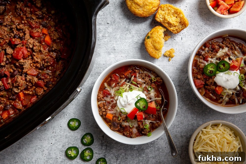 Slow-Cooker Beef Chili Recipe with Rich Smoky Flavor 4 Overhead shot of two bowls of beef chili, with a slow cooker and toppings alongside.