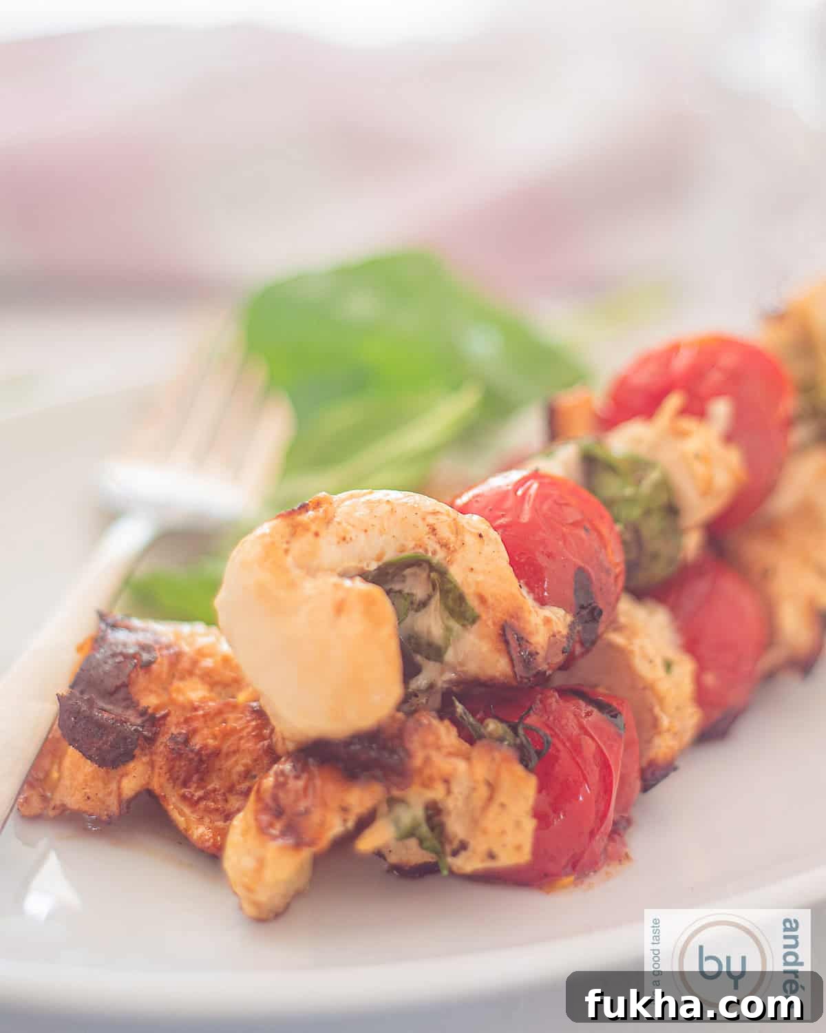 A photo of a white plate with three skewers of stuffed chicken with mozzarella and basil and grilled cherry tomatoes.