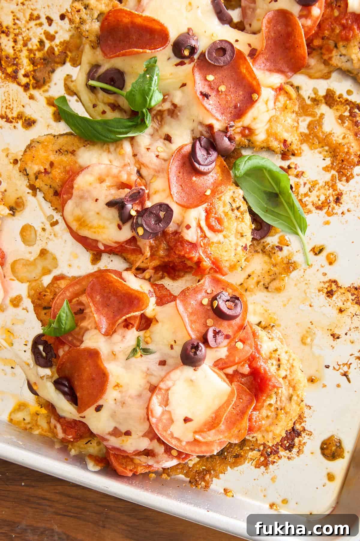 Baked chicken breasts topped with melted cheese, pepperoni slices, black olives, tomato sauce, and fresh basil leaves on a sheet pan.