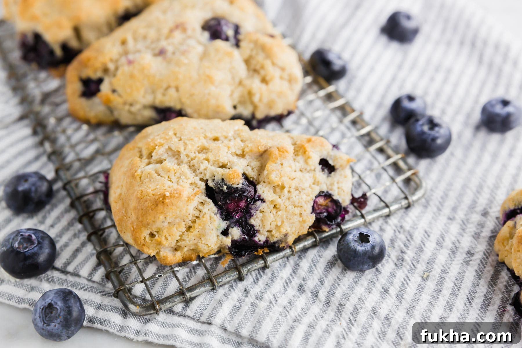 A close-up of a blueberry scone on a cooling rack, with fresh blueberries scattered on a striped cloth underneath.