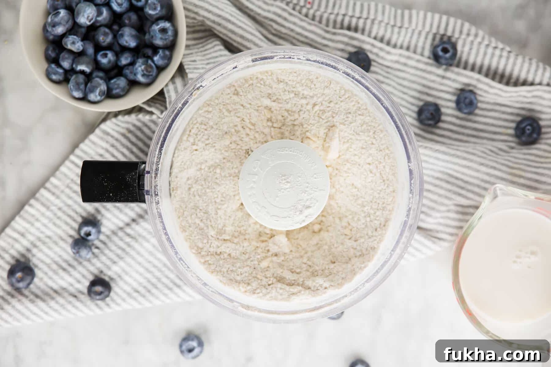 An overhead view of a food processor with flour mixture and ingredients for blueberry scones.