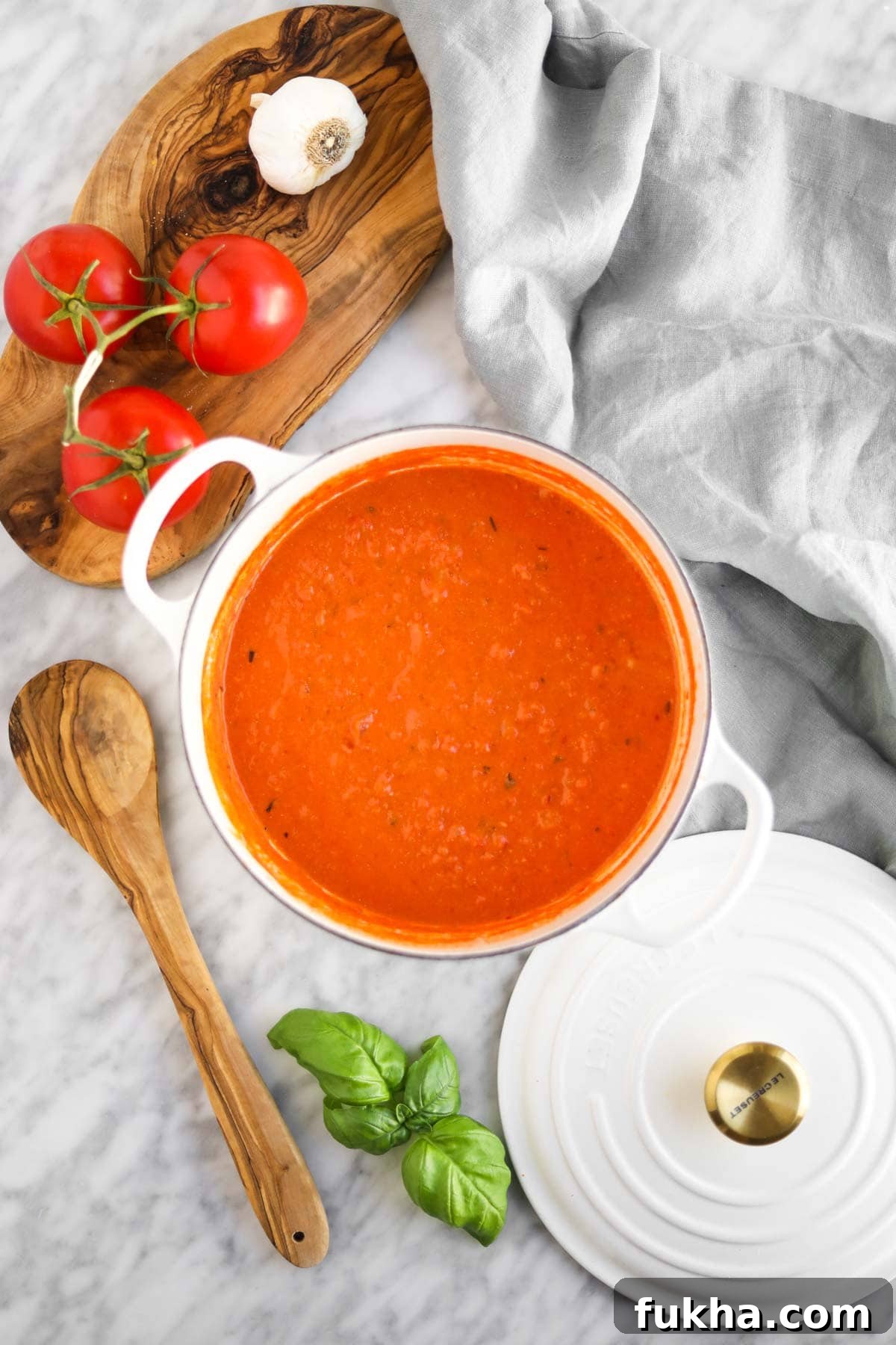 Vodka sauce in white dutch oven with wooden spoon, tomatoes, garlic, and cutting board in background.