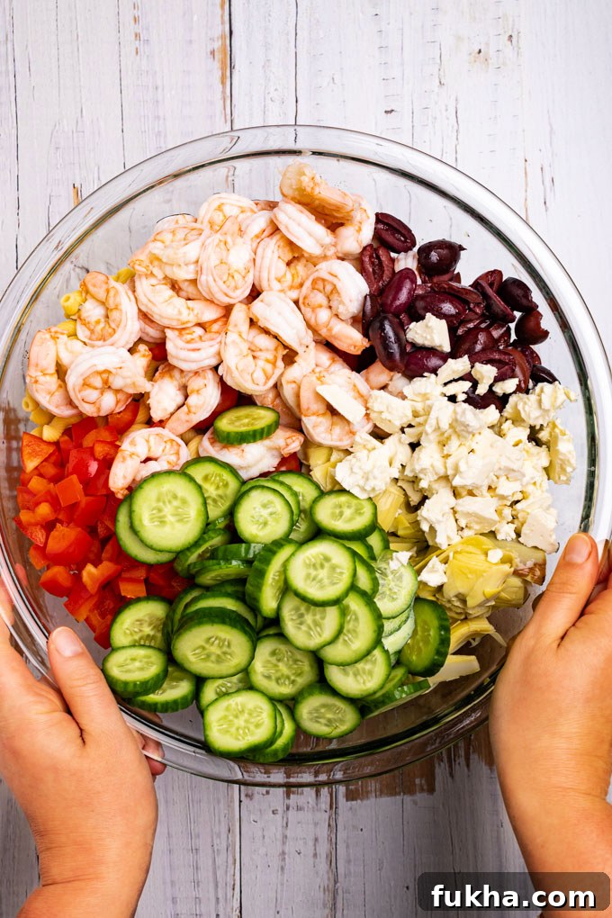 A mixing bowl of Greek pasta salad ingredients in groupings. Cucumbers, feta cheese, shrimp, kalamata olives. Hands are holding the bowl.