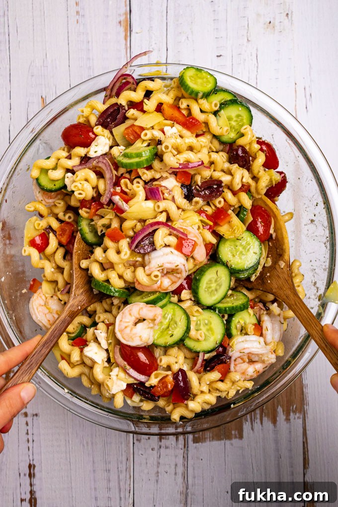 Greek pasta salad with shrimp in a mixing bowl being tossed with two wooden spoons.