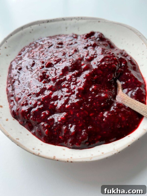A close-up of a white, speckled ceramic bowl filled with thick, dark red raspberry chia jam. A small wooden spoon rests in the jam.