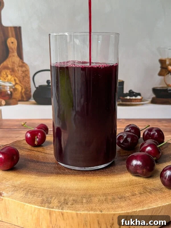 A glass is being filled with dark red cherry juice, placed on a wooden board with several fresh cherries around it.