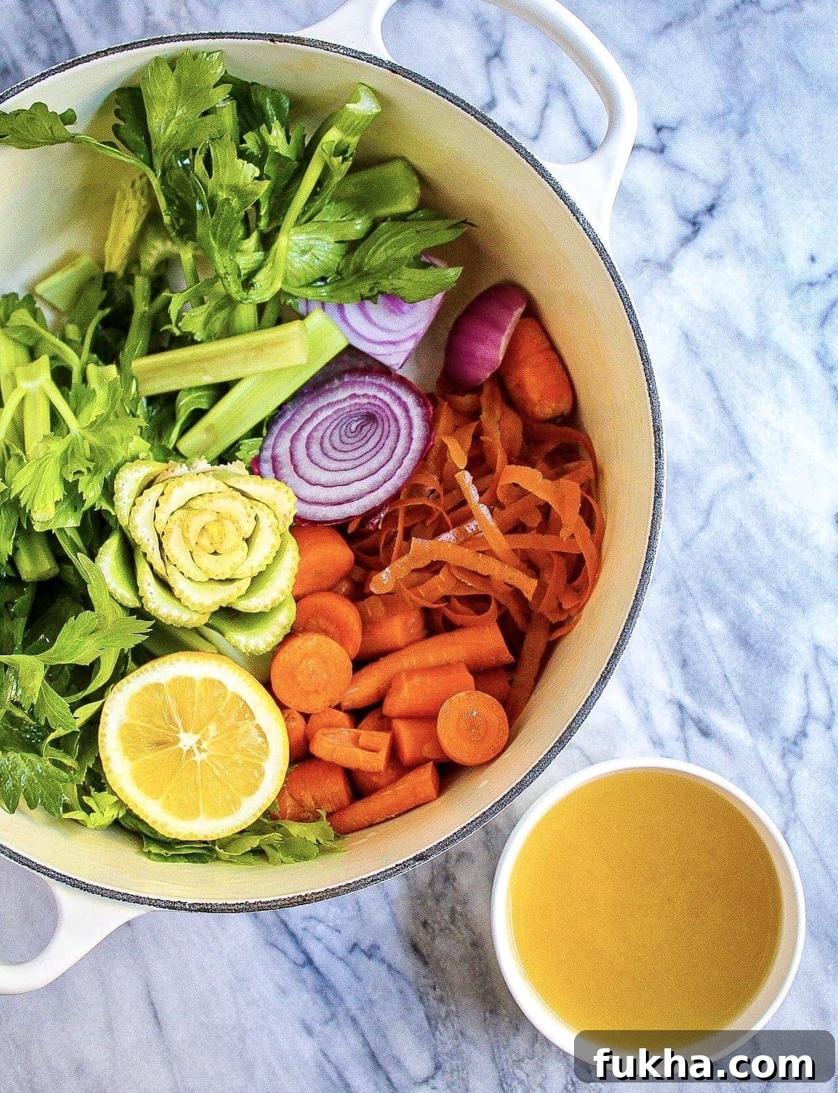 A large pot filled with celery, carrots, onion, lemon, and a small white bowl filled with broth next to it.