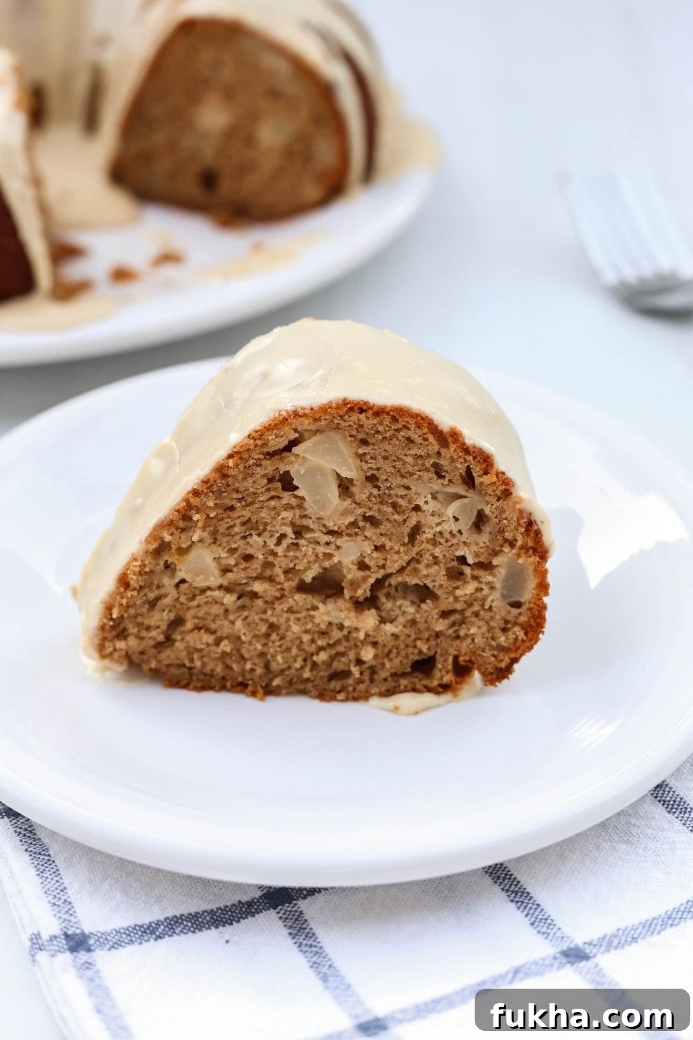 side view close-up of a slice of iced pear cake from a mix, served on a white plate, with the remaining bundt cake in the background.