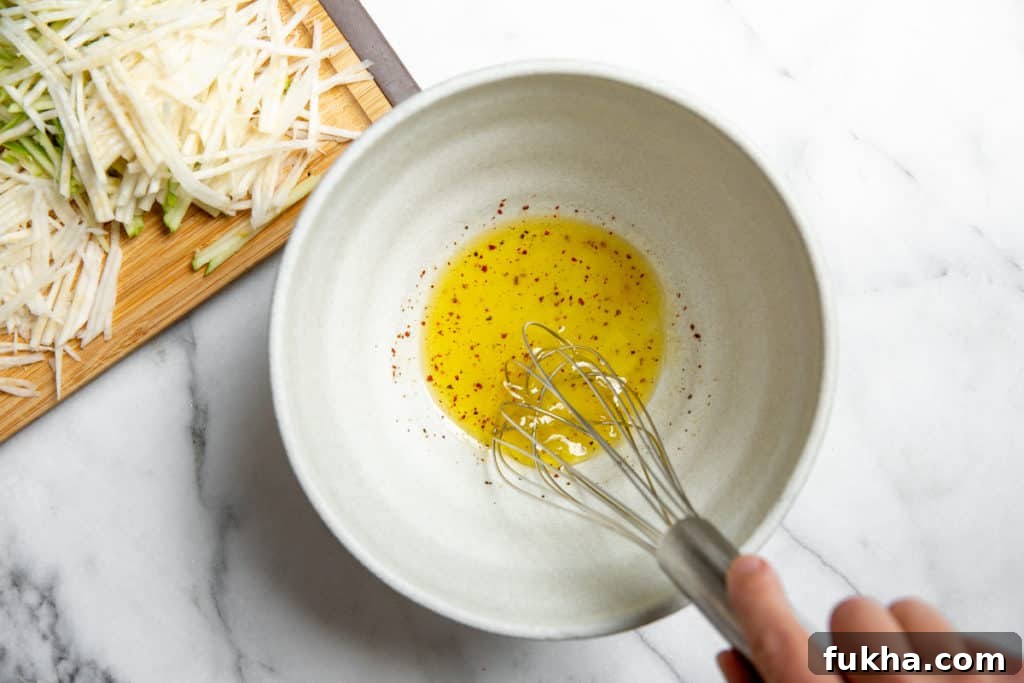 A hand whisking the lemon vinaigrette for the no-mayo apple slaw in a serving bowl.