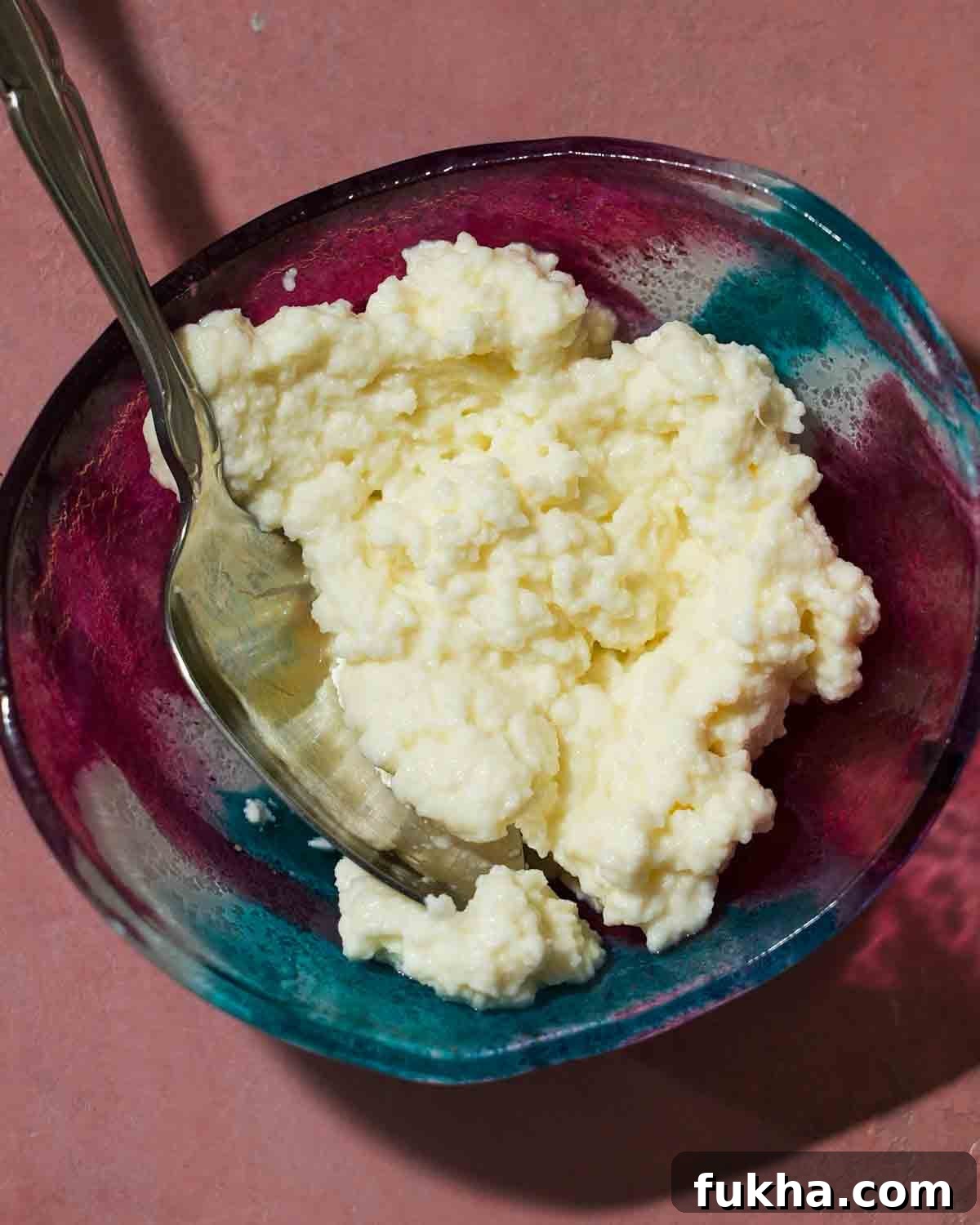 Homemade ricotta in a colorful bowl with a spoon on a pink table.