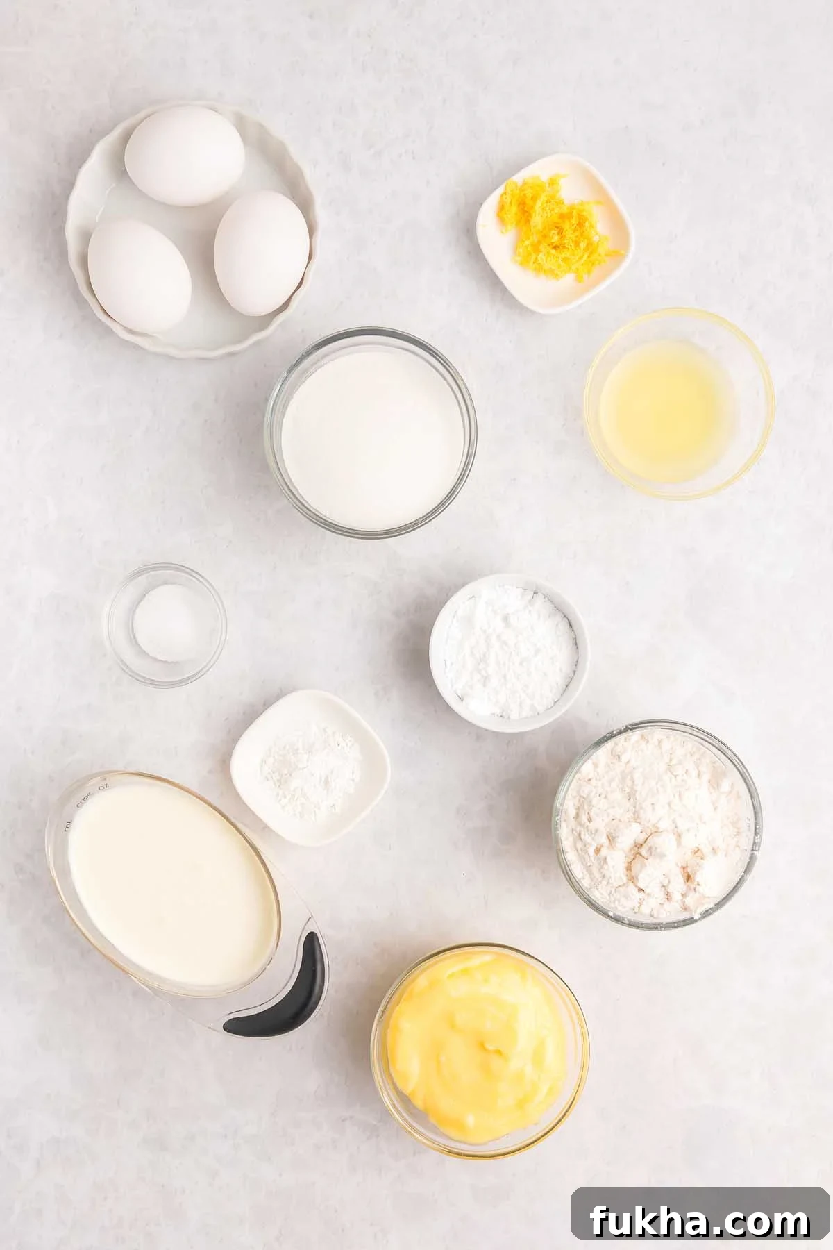 Zesty Lemon Swiss Roll Cake Recipe 13 Baking ingredients in bowls for a lemon roll cake: eggs, sugar, lemon zest, lemon juice, flour, cream, butter, and powders are arranged on a white surface.