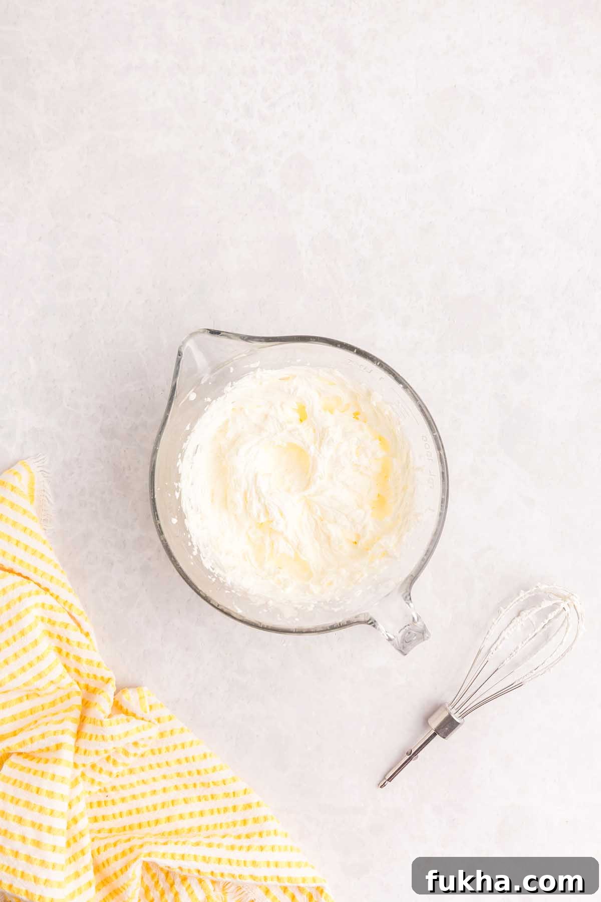 Zesty Lemon Swiss Roll Cake Recipe 16 Whipped cream in a glass bowl with a whisk and a yellow striped towel.