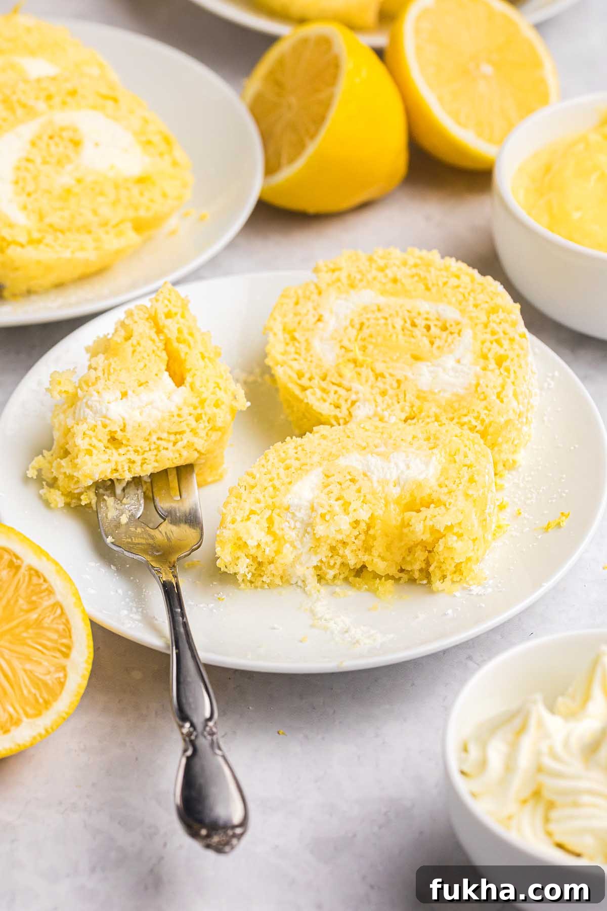 Zesty Lemon Swiss Roll Cake Recipe 4 Slices of lemon Swiss roll cake with cream filling rest on a white plate, surrounded by fresh lemons and dollops of cream.