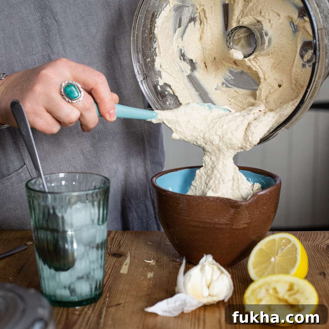 Womans hand scraping hummus from a food processor bowl into a small serving bowl