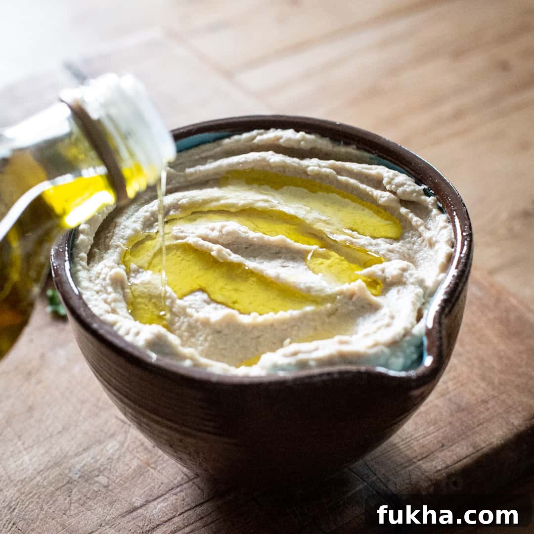 Close up shot of ceramic bowl of butter bean hummus being topped with olive oil poured form a glass bottle