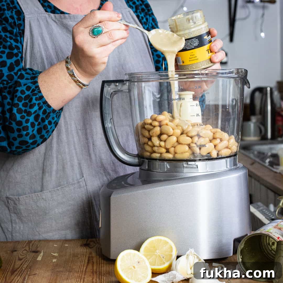 Woman in grey pinky pouring tahini from a silver spoon into a food mixer