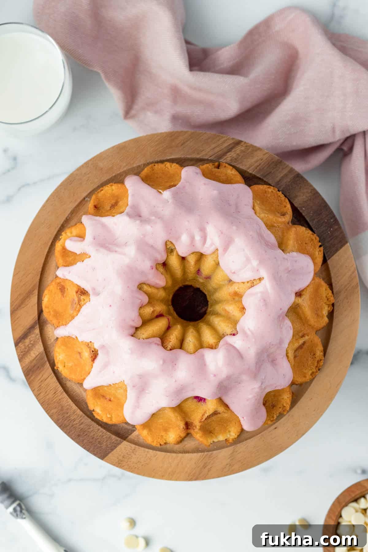 Decadent White Chocolate Raspberry Bundt Cake Recipe 2 white chocolate raspberry bundt cake on a wood cake stand with raspberry cream cheese glaze and a pink linen