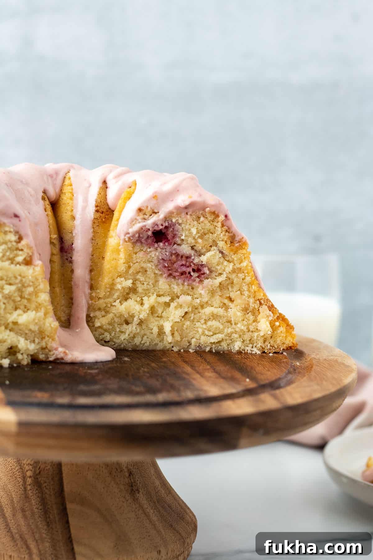 Decadent White Chocolate Raspberry Bundt Cake Recipe 3 bundt cake with pink glaze on wooden pedestal with slice exposed and glass of milk in background