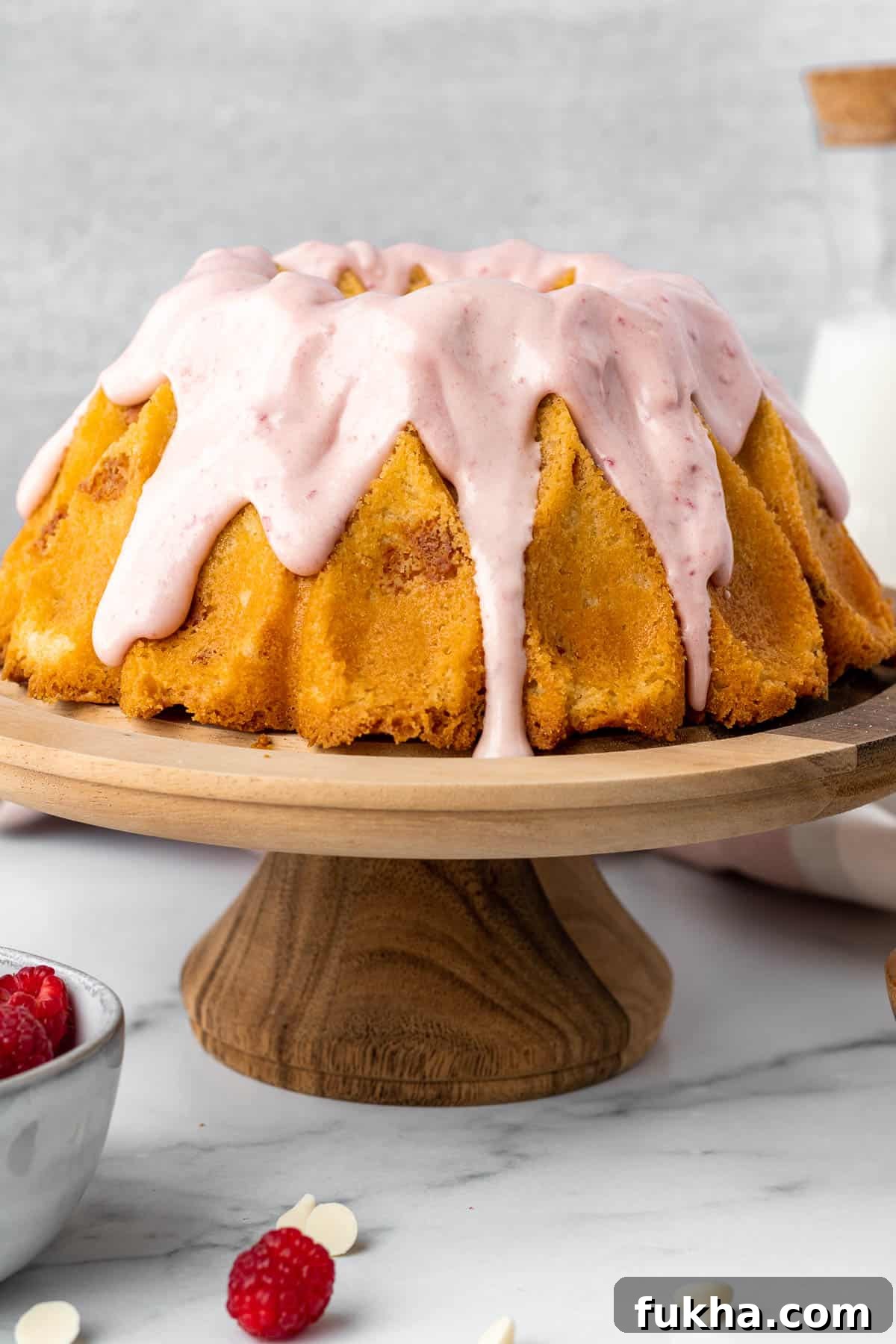 Decadent White Chocolate Raspberry Bundt Cake Recipe 4 white chocolate raspberry bundt cake on a wood pedestal with bowl of raspberries in foreground and glass of milk in background
