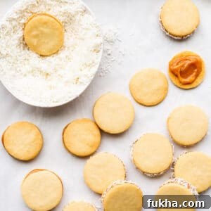 Classic Argentinian Alfajores Recipe with Dulce de Leche 13 Alfajores cookies filled with dulce de leche being rolled in a bowl of shredded coconut to coat the edges.