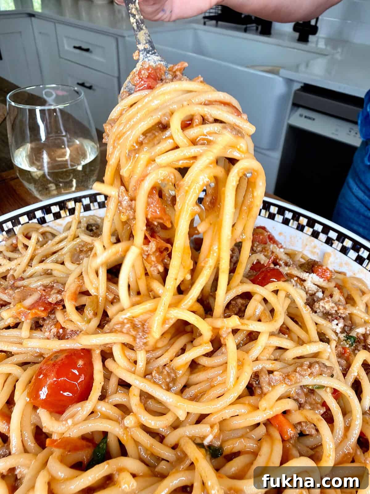 A close-up of a fork lifting Summer Spaghetti Bolognese with tomato and meat sauce from a patterned plate, with a glass of white wine and a kitchen in the background.