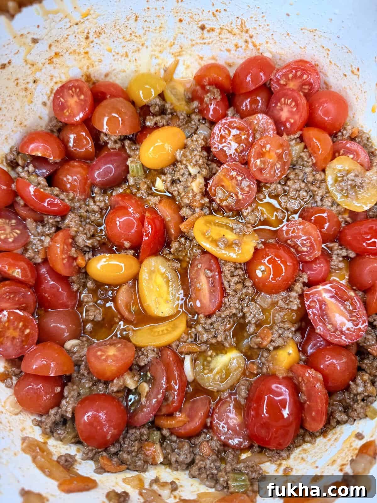 A close-up of halved red and yellow cherry tomatoes mixed with cooked ground beef and juices in a white bowl.