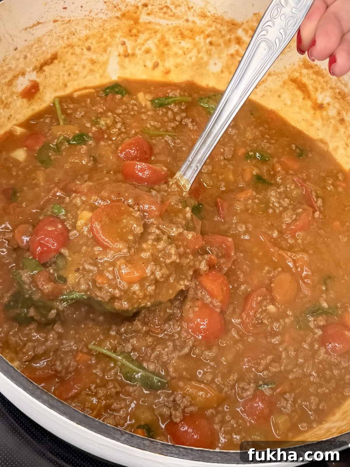 A close-up of a pot of hearty tomato-based meat sauce with ground beef, cherry tomatoes, and spinach, being stirred with a silver ladle by a person with red-painted nails.