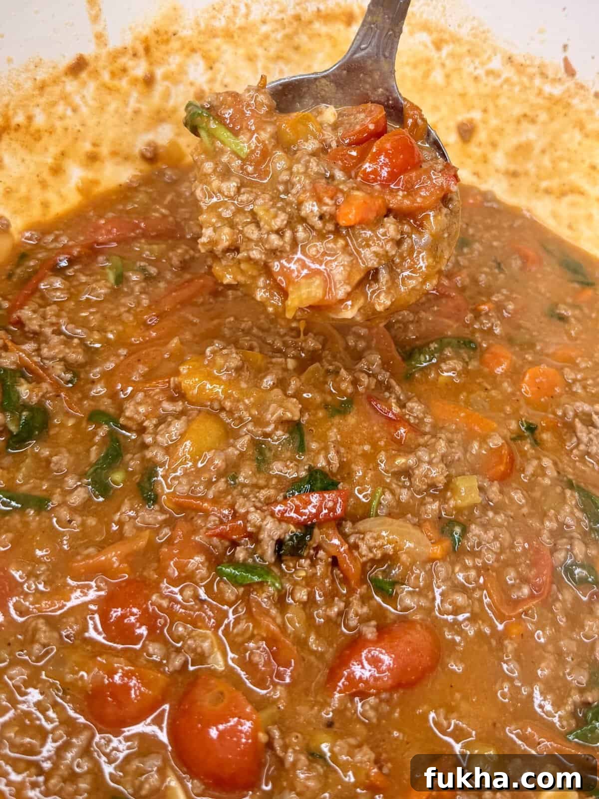 A close-up of a spoonful of hearty chili with ground beef, halved cherry tomatoes, spinach, and a rich, reddish-brown sauce in a pot.