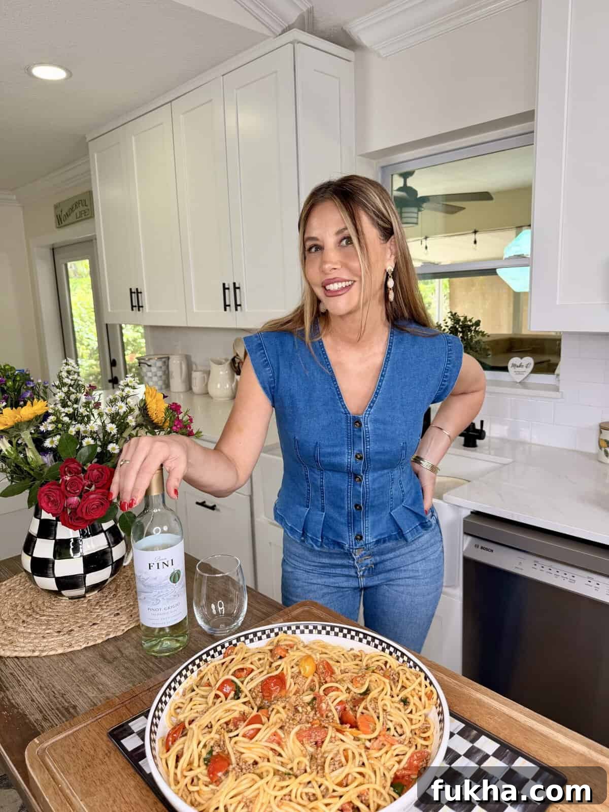 A woman in a blue denim top stands in a bright kitchen, smiling and holding a bottle of white wine next to a glass. In front of her is a large plate of pasta with tomatoes. A bouquet of flowers sits on the counter.