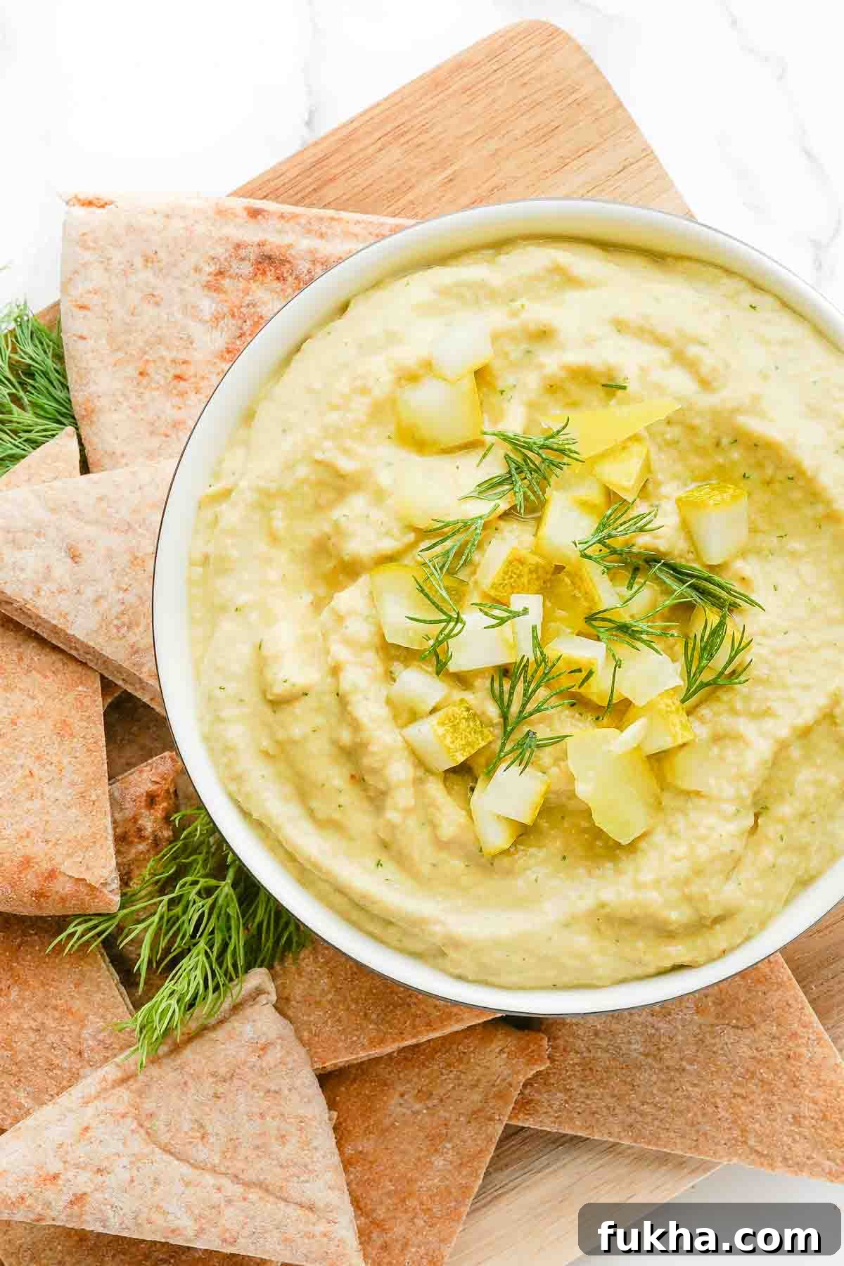 bowl of hummus garnished with pickles and dill with pita on the side.