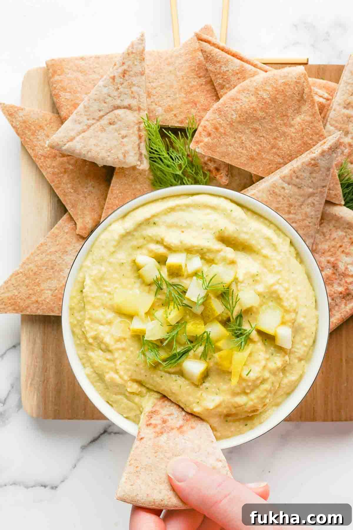 hand dipping pita triangle into hummus garnished with dill and chunks of pickle with pitas on the side.
