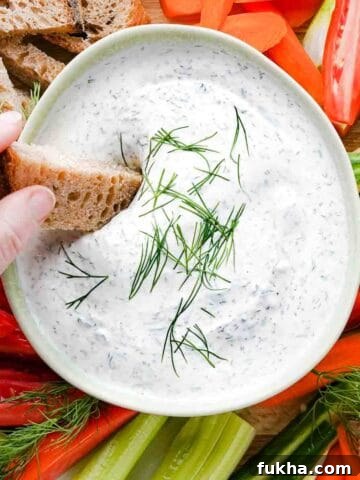 hand dipping slice of bread into yogurt dill dip garnished with dill with veggies on the side.