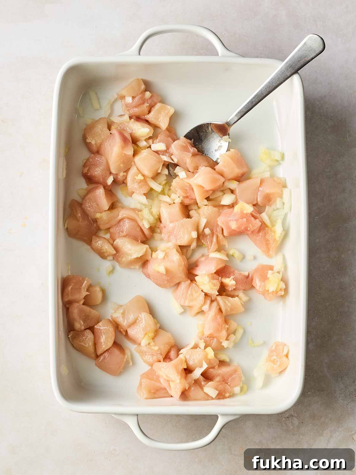 Dump-and-Bake Chicken Kyiv with Creamy Orzo 5 Cubed raw chicken in an oven dish with onion and a spoon.