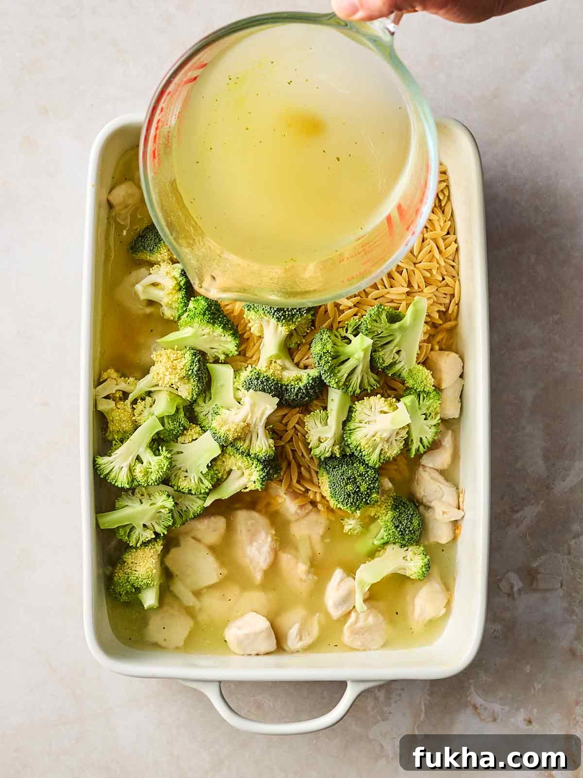 Dump-and-Bake Chicken Kyiv with Creamy Orzo 6 An oven dish with chicken, orzo, broccoli and a hand pouring in chicken stock from a jug.
