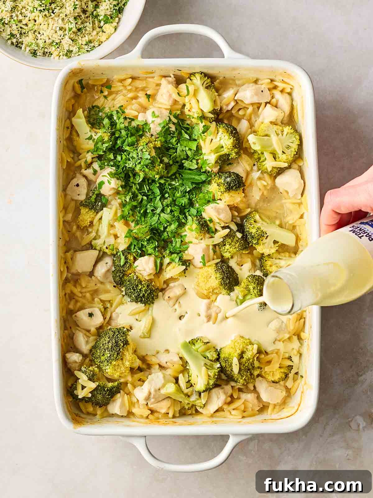 Dump-and-Bake Chicken Kyiv with Creamy Orzo 8 An oven dish filled with chicken, orzo pasta, broccoli, with a hand pouring in some double cream.