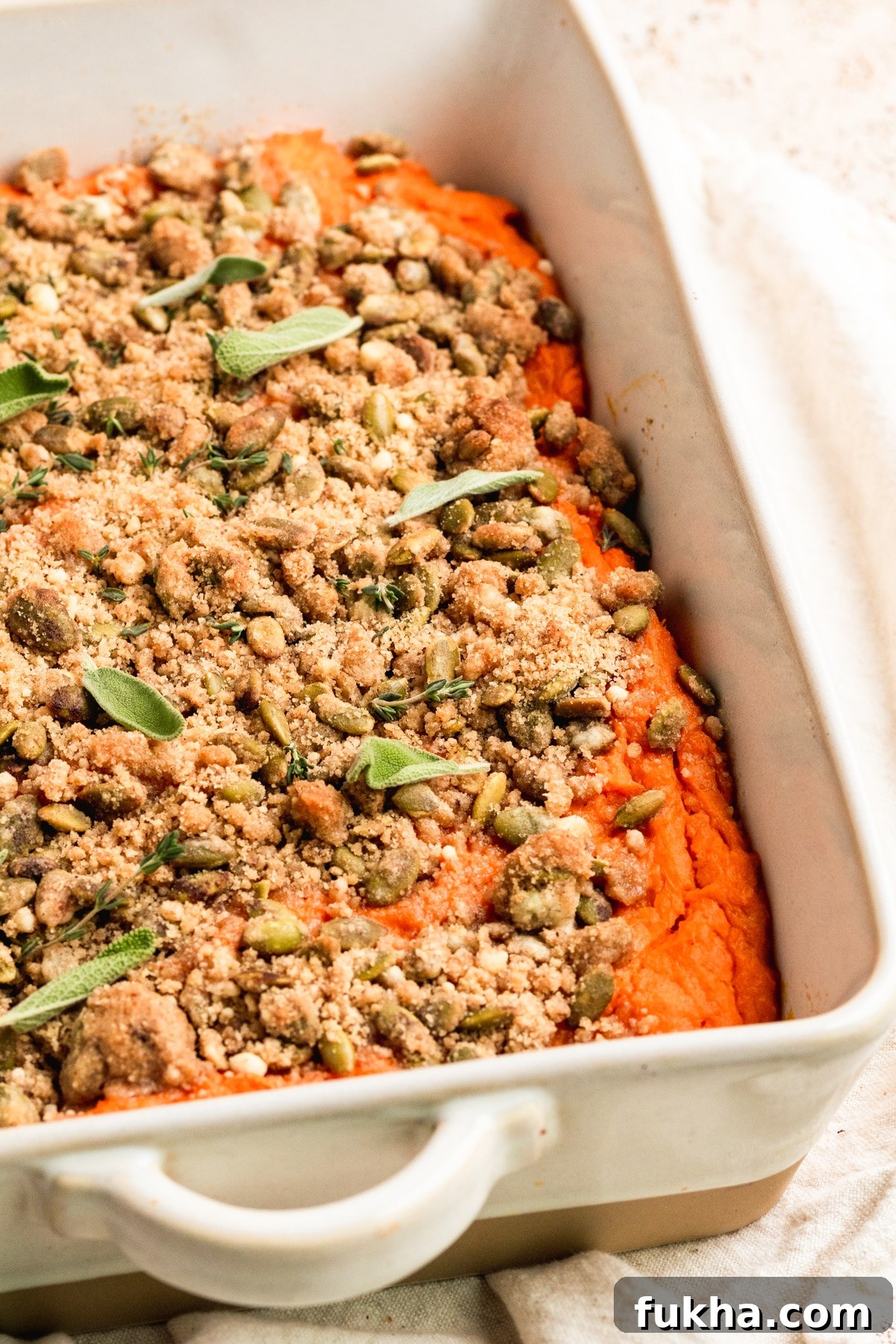 sweet potatoes in a white casserole dish topped with a brown sugar pepitas topping and fresh sage and fresh thyme. 