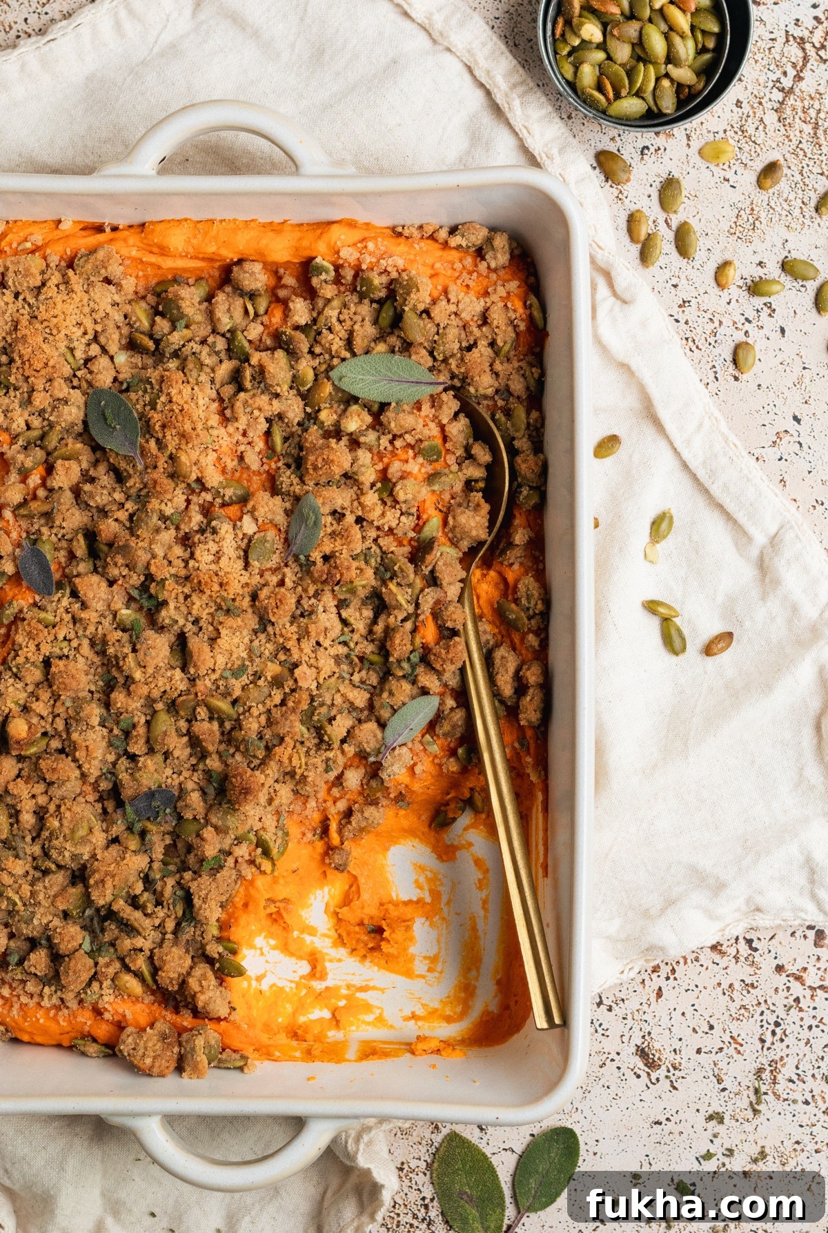 sweet potatoes in a white casserole dish topped with a brown sugar pepitas topping and fresh sage and fresh thyme with a gold spoon. 