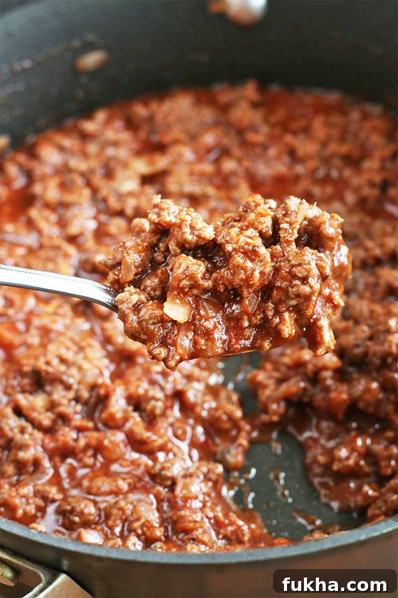 Upgraded Classic Beef Sloppy Joes Recipe 11 A large spoonful of ground beef with a red sauce is being held up to the camera, with a large stainless steel frying pan containing more of the mixture in the background.