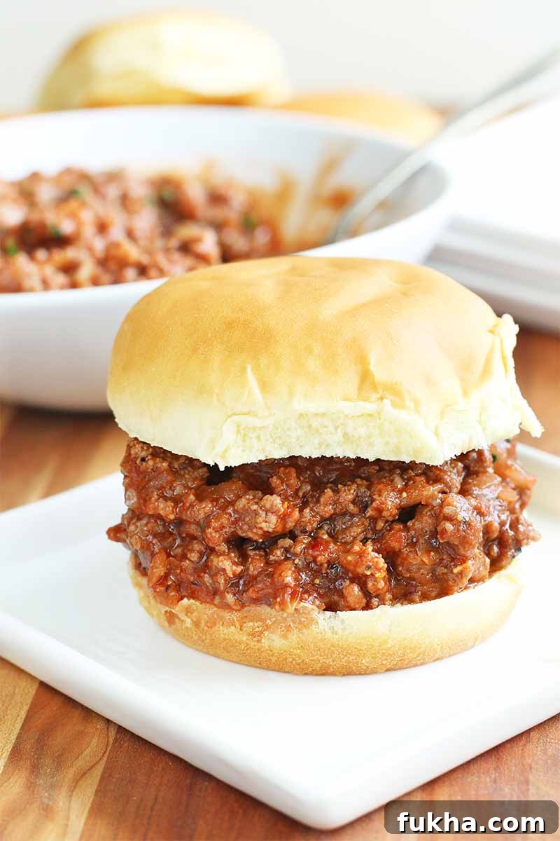 Upgraded Classic Beef Sloppy Joes Recipe 3 Homemade sloppy joe mixture on a hamburger bun, on a square white plate with more in a white bowl with a spoon in the background, on a brown wood surface.