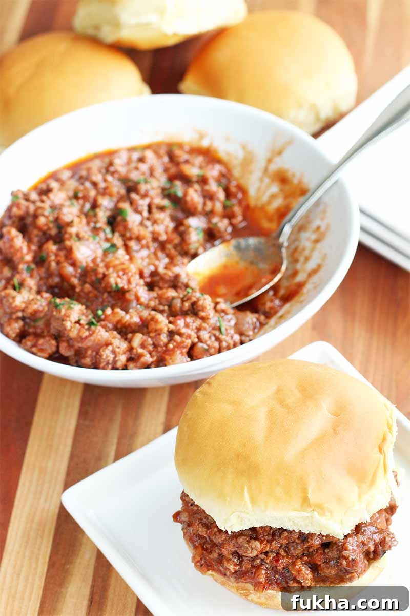 Upgraded Classic Beef Sloppy Joes Recipe 4 A large white bowl of beef sloppy joe with a stainless steel serving spoon, beside a small stack of square white plates, with a serving on a hamburger bun in the foreground, on a brown wood table.