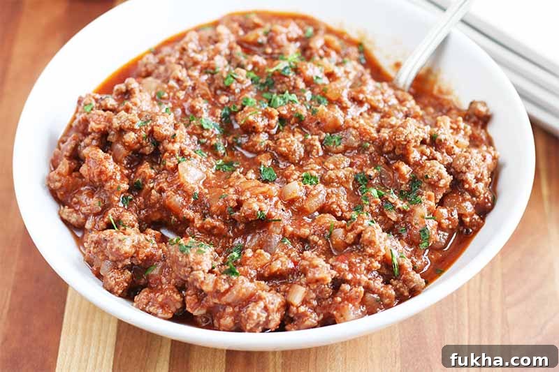 Upgraded Classic Beef Sloppy Joes Recipe 5 A bowl of homemade sloppy joe sandwich filling topped with chopped parsley, on a brown wood table beside a stack of white plates.