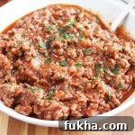 A bowl of homemade sloppy joe sandwich filling topped with chopped parsley, on a brown wood table beside a stack of white plates.
