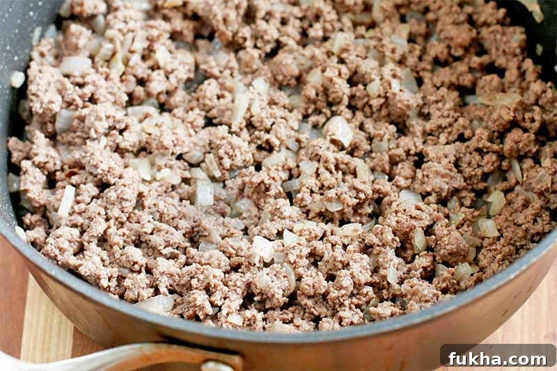 Upgraded Classic Beef Sloppy Joes Recipe 9 Ground beef browning in a large nonstick frying pan with chopped onions, on a brown wood surface.