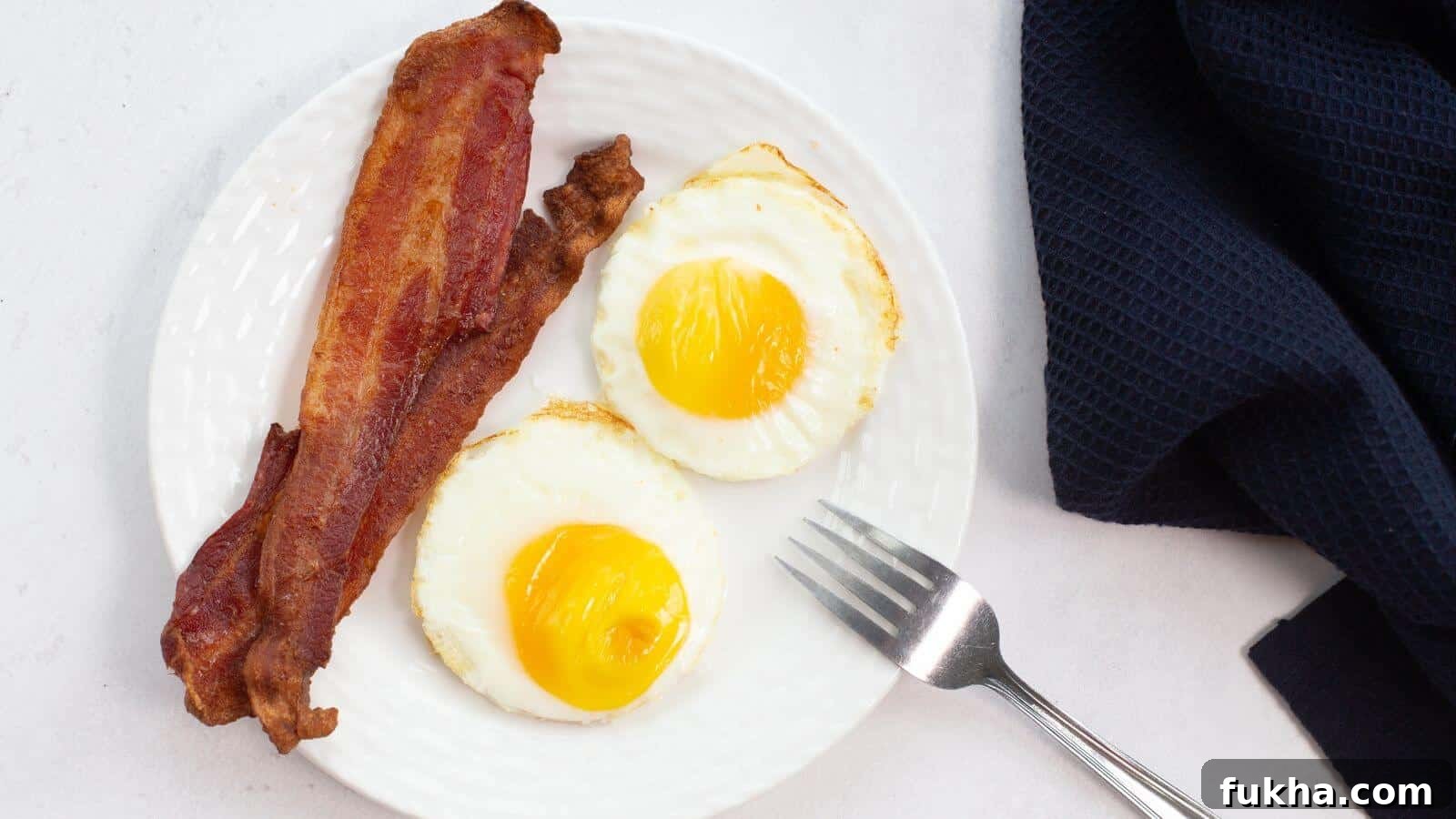 24 Low-Carb Breakfasts to Keep You Full Until Noon 15 Plate with two sunny-side-up eggs, perfectly cooked like fried eggs in an air fryer, and two strips of crispy bacon, next to a fork and a dark cloth napkin.