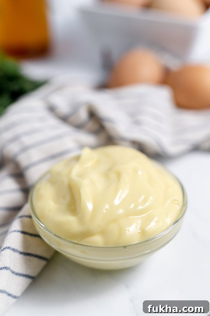 Image shows a small dish of homemade mayonnaise on a table next to a striped towel. 