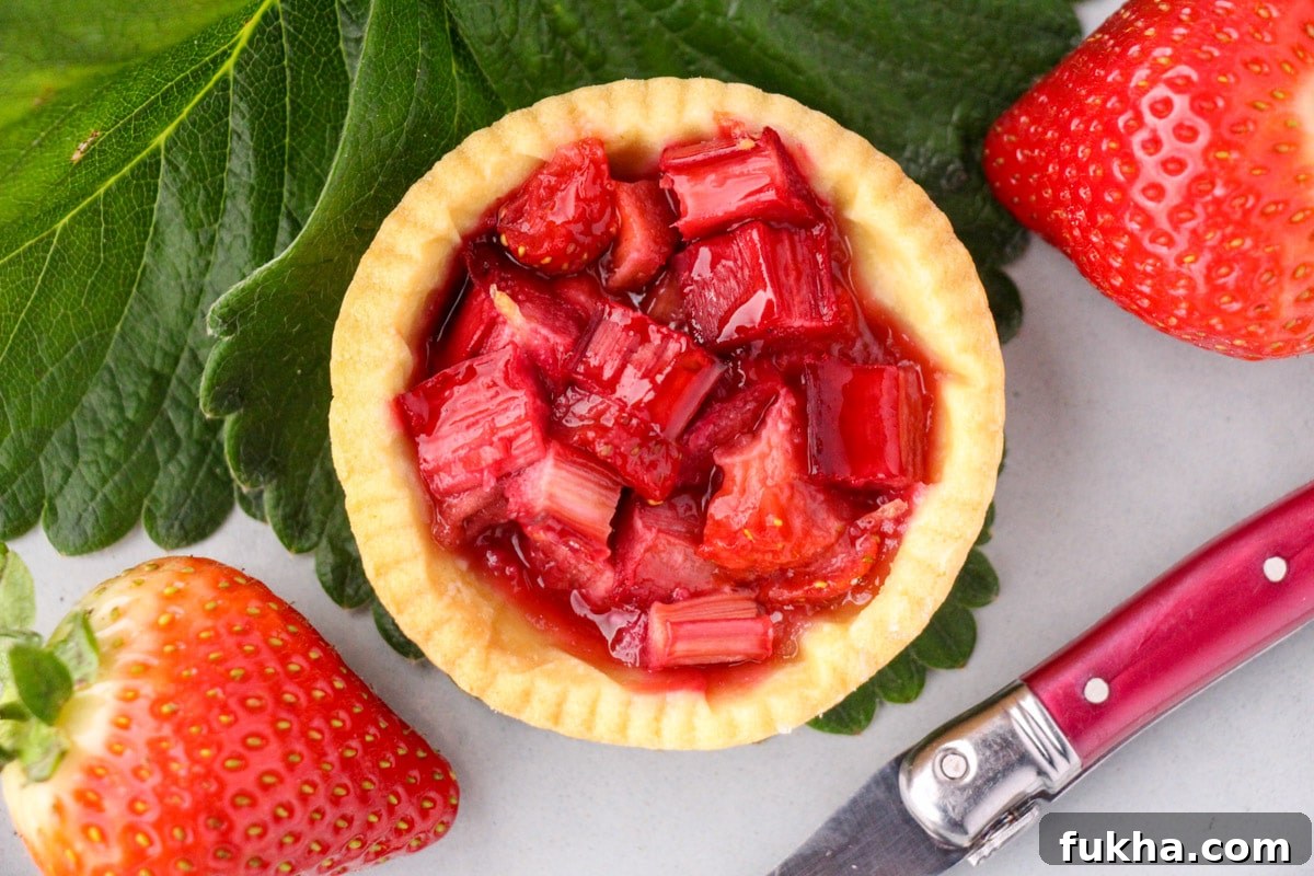 Close up strawberry rhubarb tartlets with strawberry leaves and fresh strawberries and a vintage cake fork on the side.
