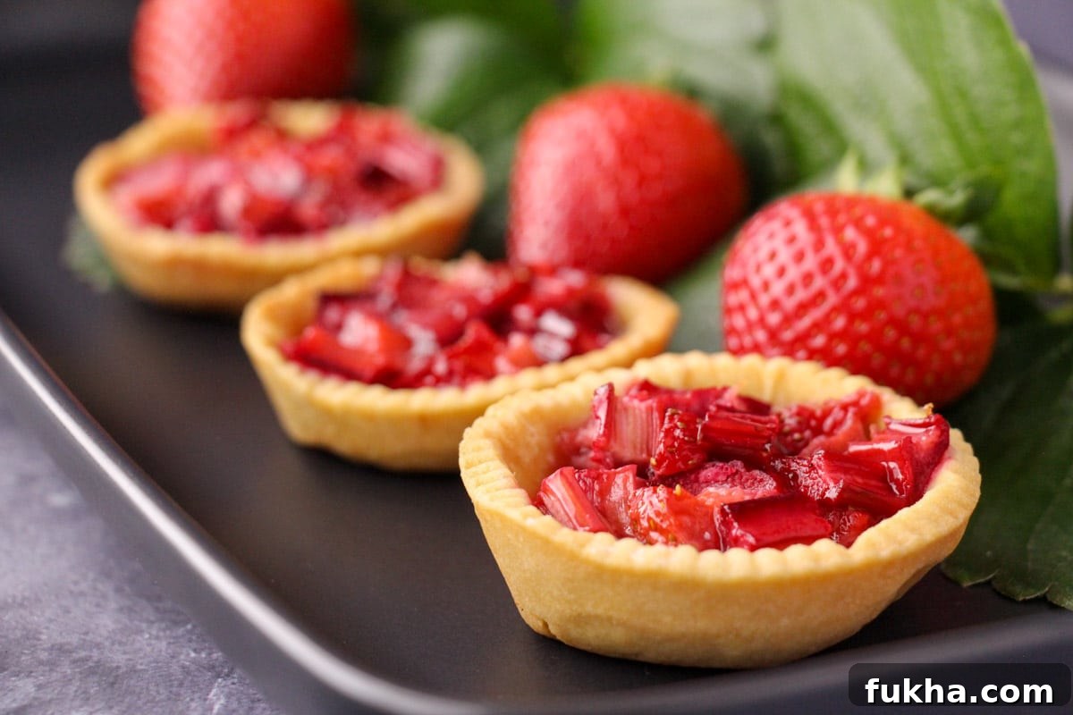Row of strawberry rhubarb tartlets on a black serving plate, with fresh strawberries and strawberry leaves on the side.